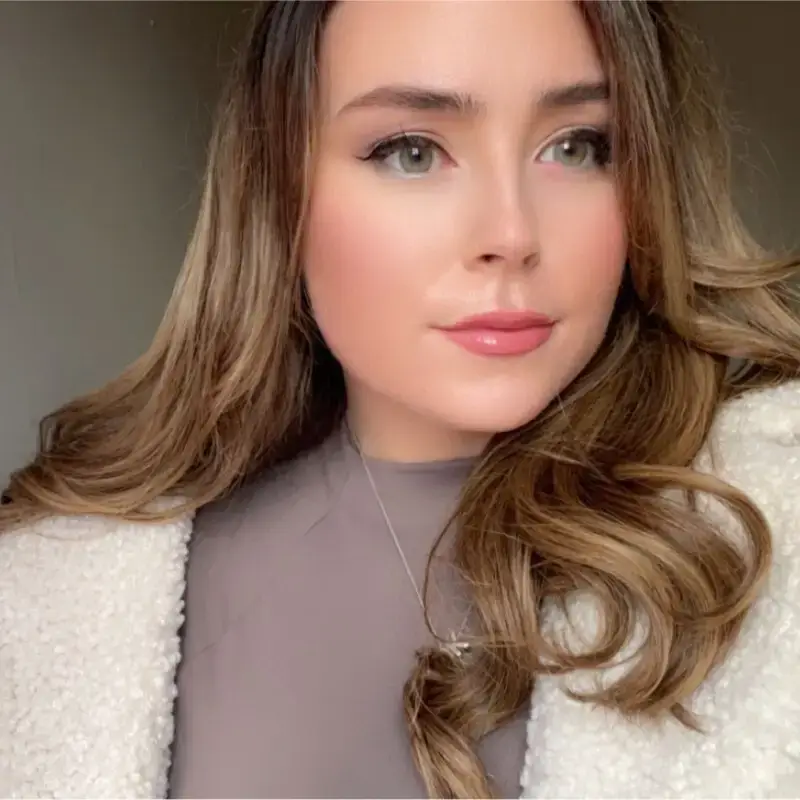 A young woman with long, wavy light brown hair looks slightly to the side. She wears a light-coloured faux fur coat over a taupe top, styled for a healthcare digital marketing agency shoot. Her make-up is natural with pink lipstick and a softly blurred background.