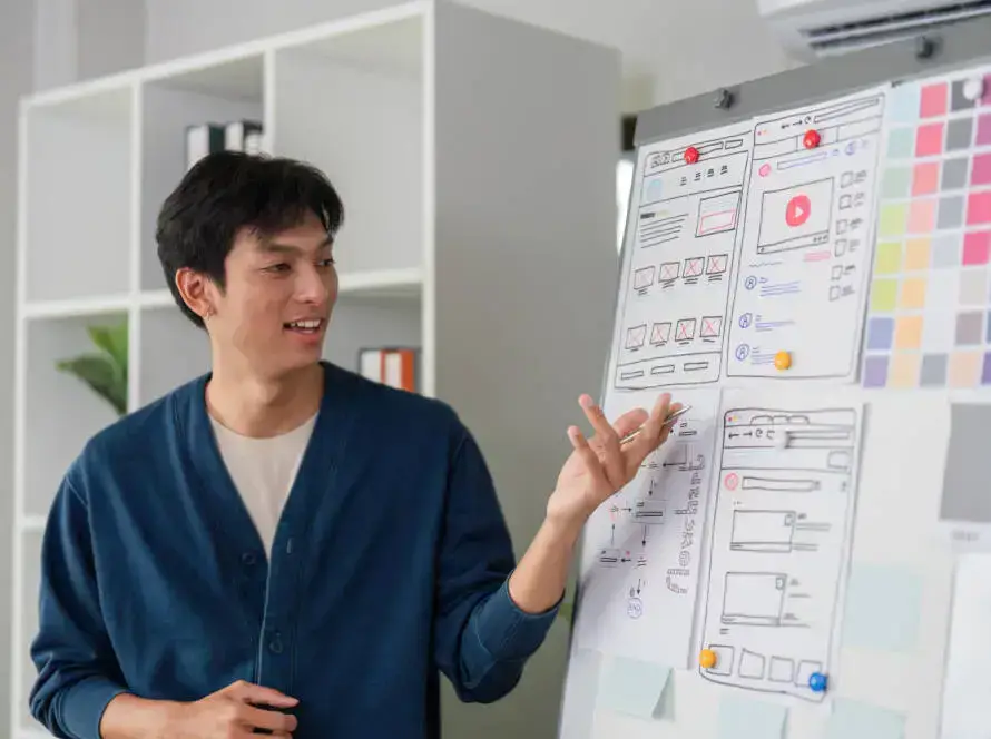 A person wearing a blue cardigan gestures towards a flip chart displaying UI/UX wireframes and website layout sketches, illustrating digital marketing healthcare strategies. White shelves with books and folders line the bright, modern office background.