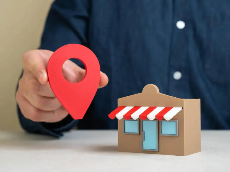 A person in a dark blue shirt holds a large red map location pin beside a small model shopfront, symbolising how a healthcare digital marketing agency can boost local visibility for clinics using expert healthcare SEO strategies.