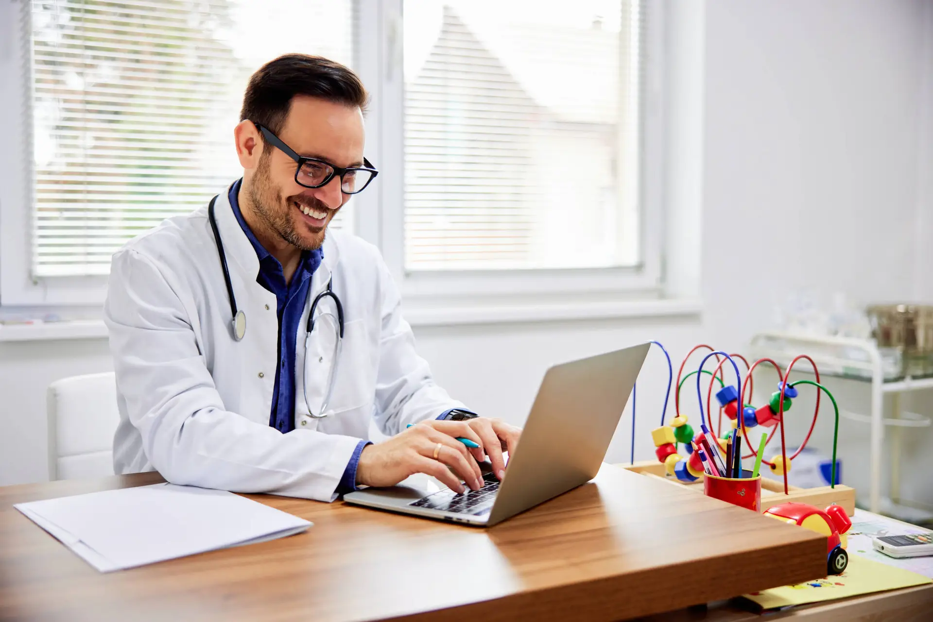 A smiling doctor in glasses and a white coat types on a laptop at a wooden desk, likely researching healthcare SEO. A stethoscope hangs around his neck, with papers and a colourful bead maze nearby. Sunlight streams through window blinds in the background.
