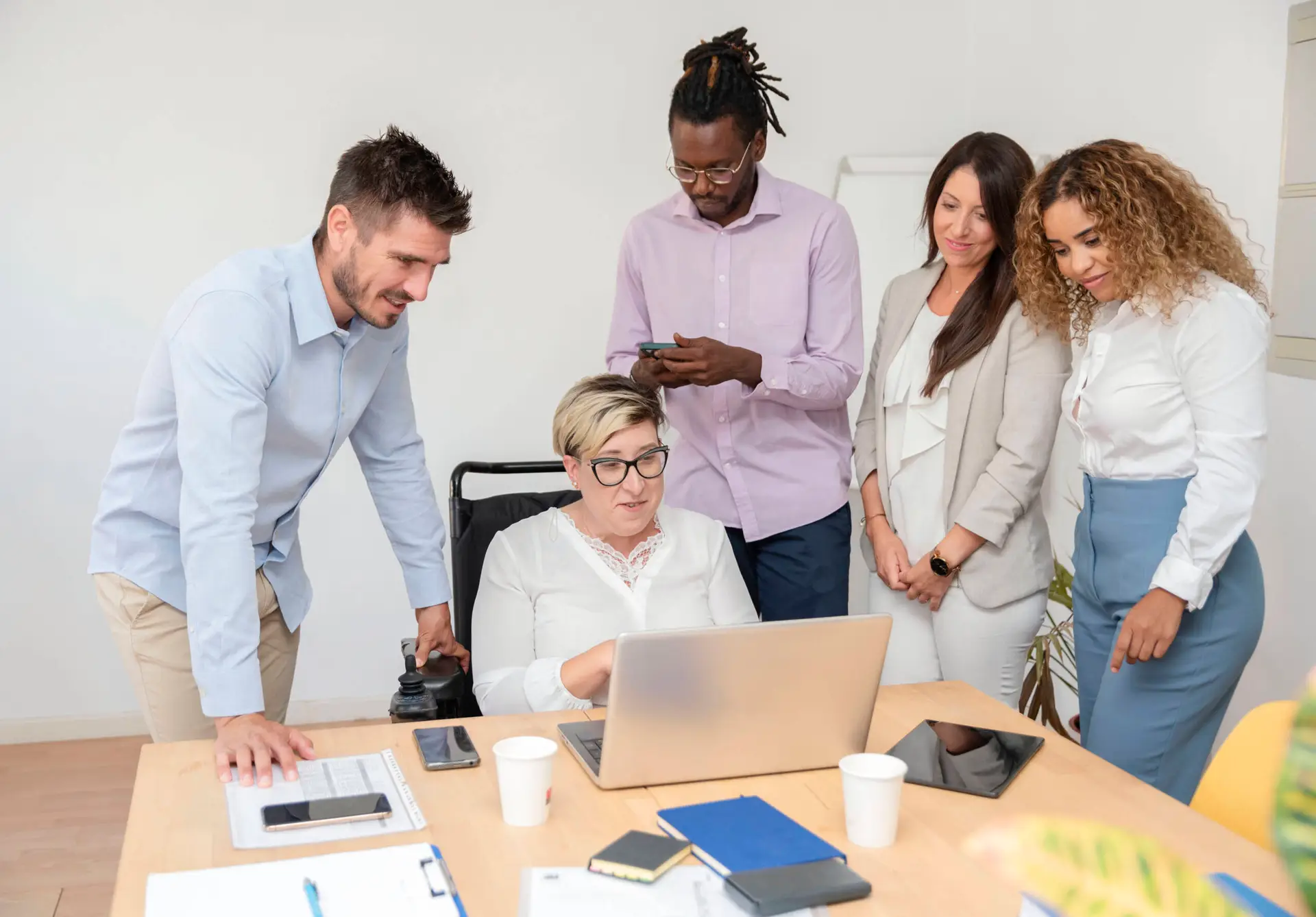 Five diverse colleagues gather around a woman in a wheelchair working on a laptop. Papers, two cups, a phone, and a tablet are on the table as this engaged team from a healthcare digital marketing agency collaborates in a bright office setting.