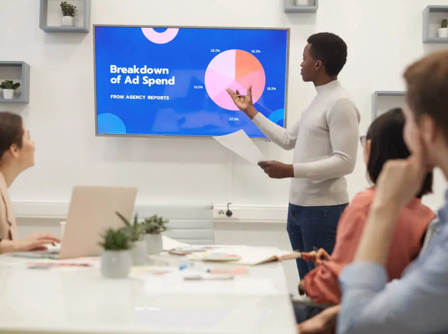 A man from a healthcare digital marketing agency presents a pie chart titled “Breakdown of Ad Spend” to three colleagues in a modern office. The group listens attentively, surrounded by laptops, plants, and papers on the table.