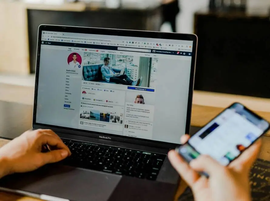 A person uses a MacBook displaying a Facebook profile page whilst holding a mobile phone. Both devices show social media content—perfect for a healthcare digital marketing agency seeking new inspiration and effective outreach strategies.