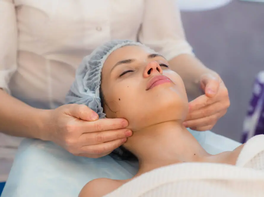 A person lies on a spa bed with eyes closed, wearing a disposable hair cap and a white towel. Another gently massages their jawline and neck, in a calm setting that could inspire visuals for a healthcare SEO consultant’s wellbeing-focused campaign.