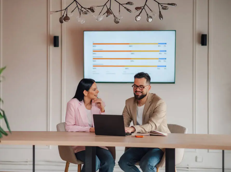 Two people sit at a desk with a laptop and notebook, smiling and talking. Both wear business attire. Behind them, a screen displays a colourful bar chart—typical of a modern healthcare digital marketing agency meeting room with upmarket décor and greenery.