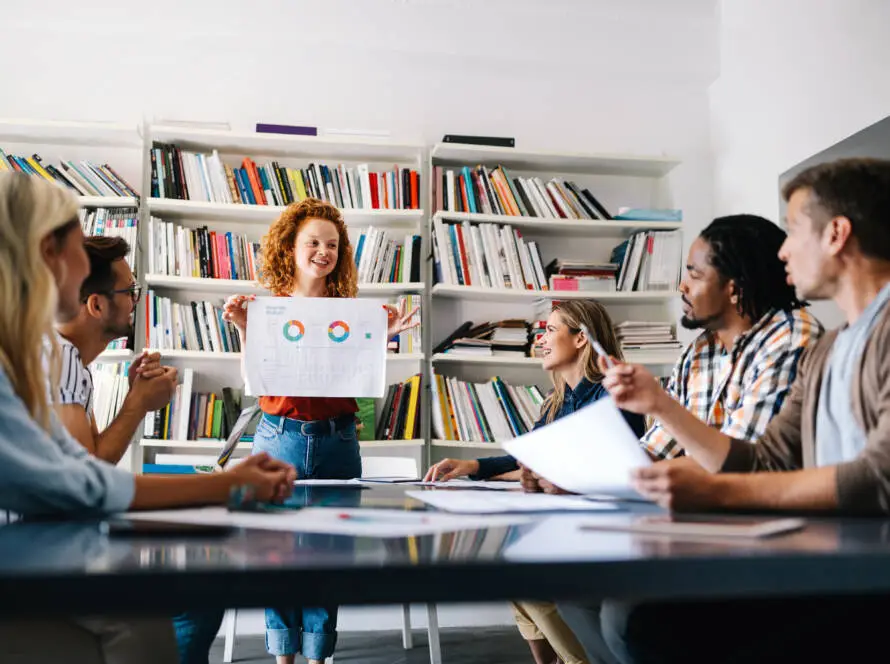 A woman with curly red hair stands in front of a bookcase, smiling and holding up a presentation chart with graphs. Five people sit around a table, suggesting a collaborative meeting on digital marketing healthcare strategies.