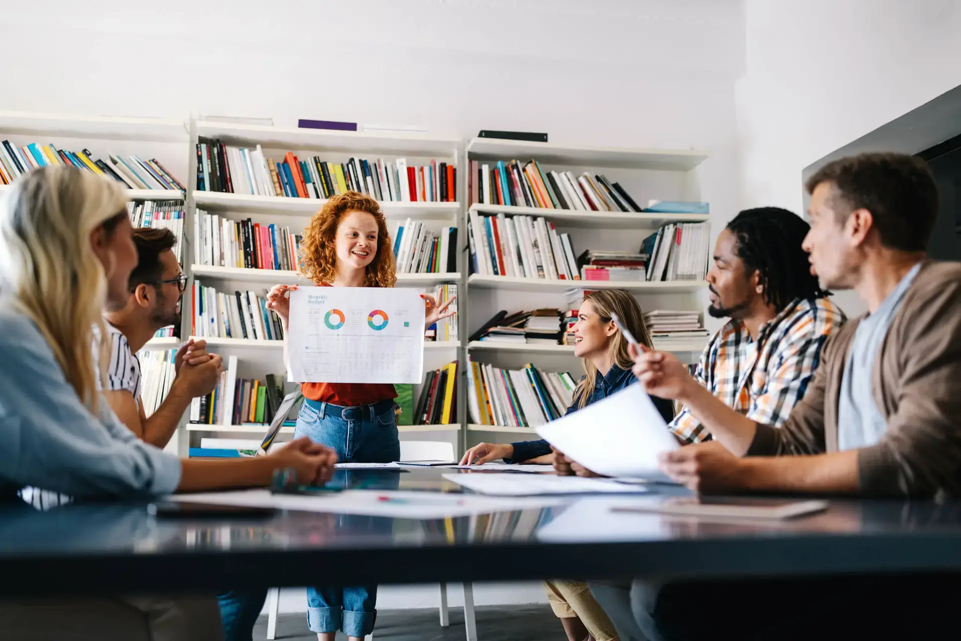 A woman with curly red hair stands in front of a bookcase, smiling and holding up a presentation chart with graphs. Five people sit around a table, suggesting a collaborative meeting on digital marketing healthcare strategies.