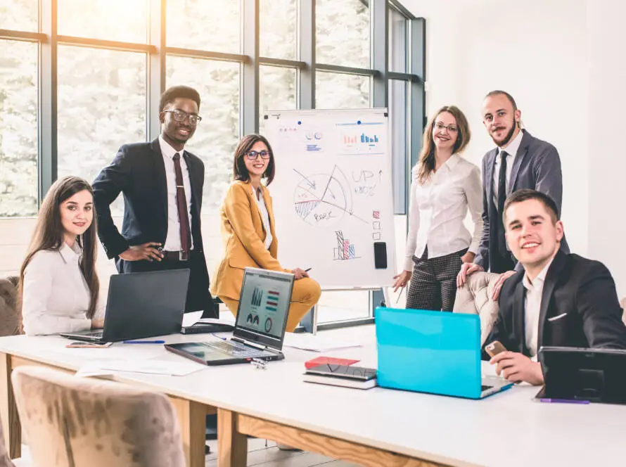 Six diverse professionals in business attire pose and smile in a bright office. Some, part of a healthcare digital marketing agency, are seated at a table with laptops, while others stand by a whiteboard with charts and graphs. Natural light adds to the collaborative atmosphere.