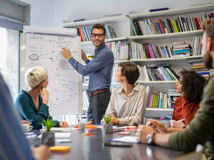 A man in glasses stands by a flipchart, pointing at diagrams while five colleagues from a healthcare digital marketing agency listen intently. Shelves filled with books line the room, setting the scene for an engaging business presentation.