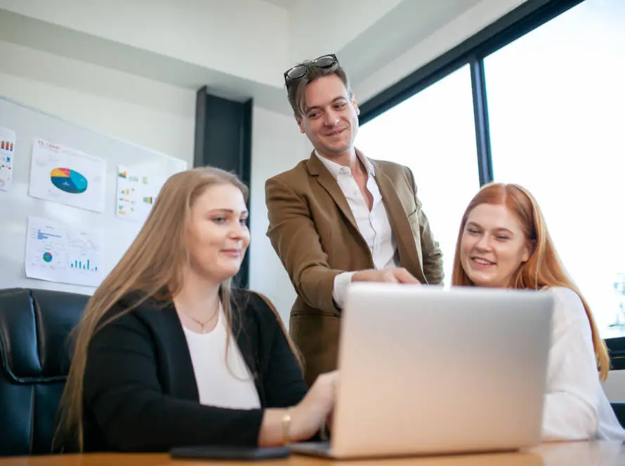 Three people in business attire gather around a laptop in a modern office. One points at the screen whilst two others sit, smiling, as they discuss strategies with a medical marketing agency. Charts and graphs are visible on the wall in the background.