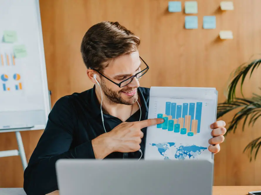 A man wearing glasses and earphones sits at a desk with a laptop, smiling and pointing to a colourful bar chart, showcasing results for a healthcare digital marketing agency. A whiteboard with graphs and sticky notes hangs on the wooden wall behind him; a plant is visible on the right.