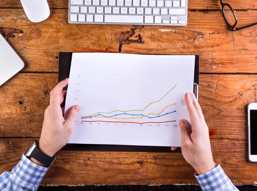 A person in a blue checked shirt holds a piece of paper with a multicoloured line graph at a wooden desk, suggesting digital marketing healthcare analysis. A keyboard, mobile phone, and paper nearby highlight an office setting tracking data trends.