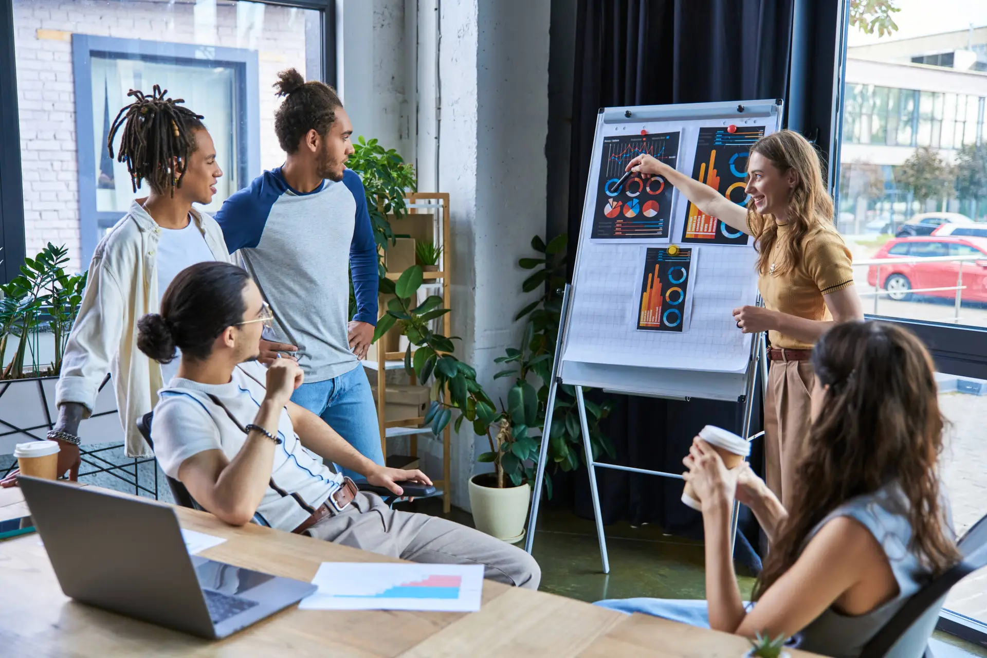 Four people gather around a table in a bright office, listening to a woman from a medical marketing agency presenting graphs and charts on a flipchart. Laptops, documents, and coffee cups are on the table, with large windows and green plants in the background.