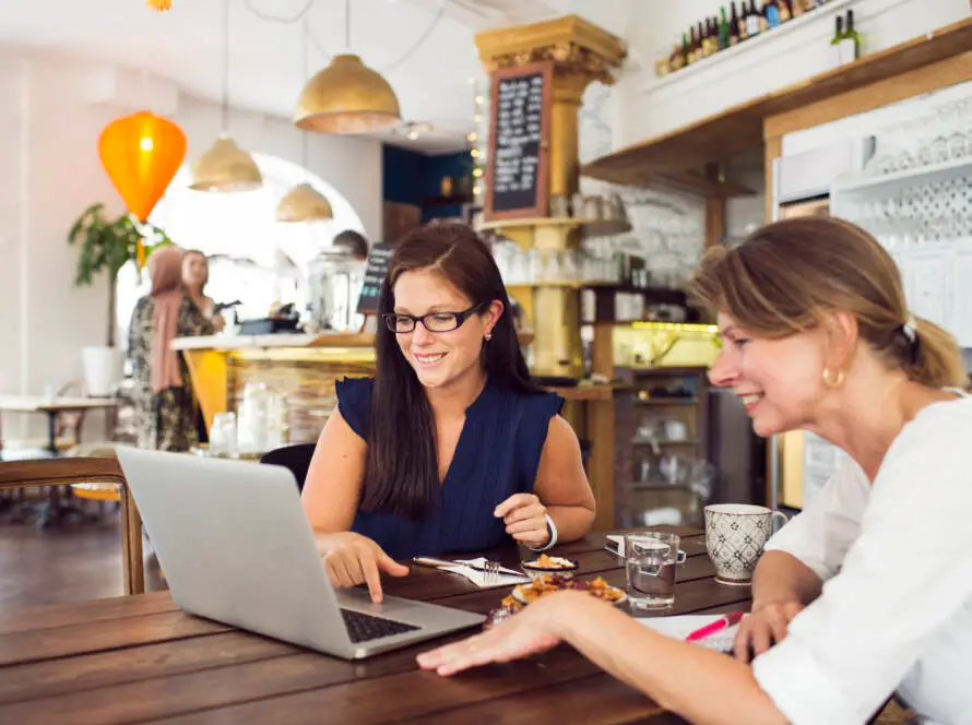 Two women sit at a wooden table in a cosy café, smiling at a laptop while discussing strategies for their healthcare digital marketing agency. Plates with food, coffee, and water are on the table, set against warm lighting and shelves in the background.