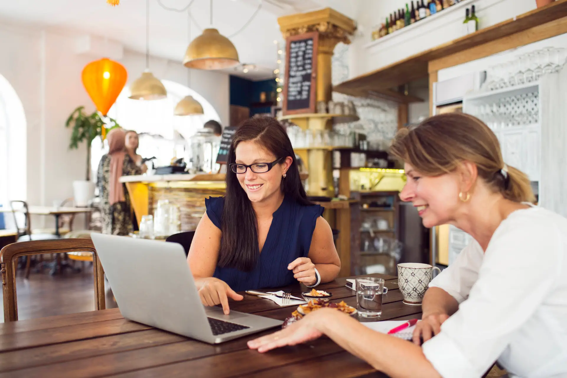 Two women sit at a wooden table in a cosy café, smiling at a laptop while discussing strategies for their healthcare digital marketing agency. Plates with food, coffee, and water are on the table, set against warm lighting and shelves in the background.