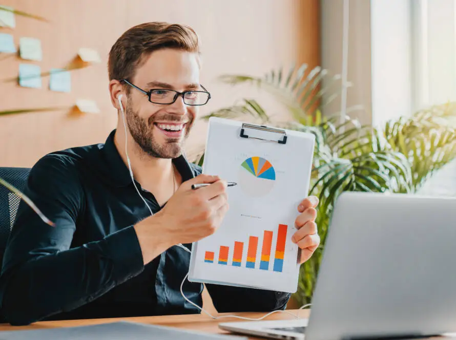 A smiling man with glasses and earphones sits at a desk, holding up a clipboard with colourful charts. He gestures at the graphs with a pen during a video call in a bright office, representing a healthcare digital marketing agency.