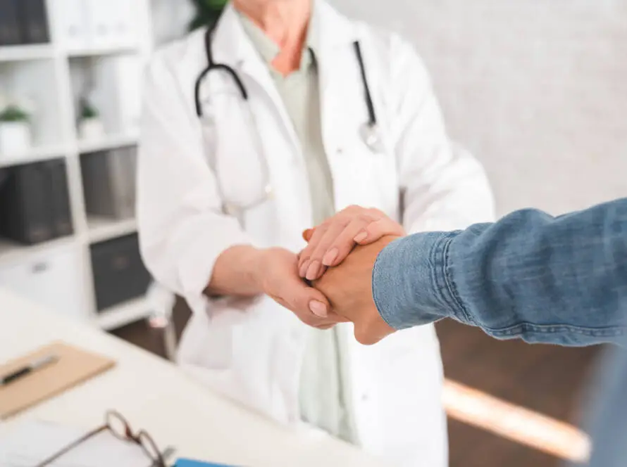 A doctor in a white coat with a stethoscope gently holds the hand of a patient across a desk, showing support and empathy—a scene that digital marketing healthcare experts often highlight to convey trust and care in bright medical surgery settings.