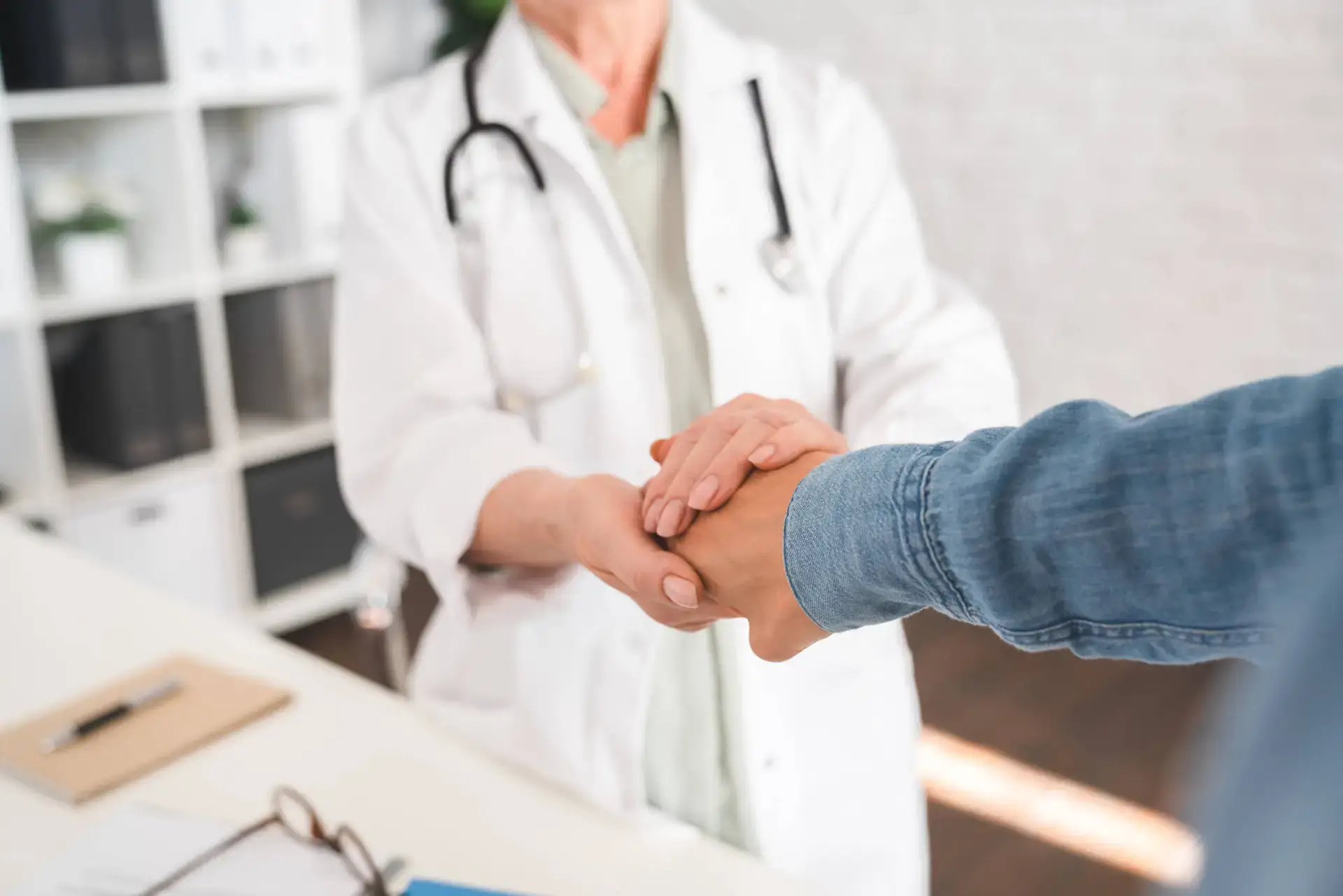 A doctor in a white coat with a stethoscope gently holds the hand of a patient across a desk, showing support and empathy—a scene that digital marketing healthcare experts often highlight to convey trust and care in bright medical surgery settings.
