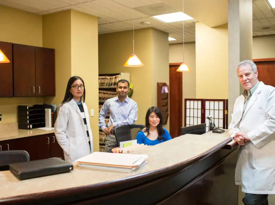 Four medical professionals stand and sit behind a curved reception desk in a clinic. As they work, their digital marketing healthcare strategies ensure the clinic stays connected with patients. The background features file shelves, cupboards, and warm lighting.