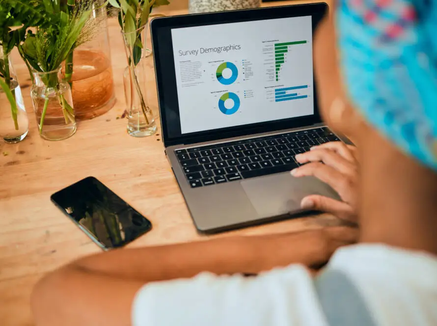 A person sits at a wooden table using a laptop displaying colourful survey demographic charts and statistics—perhaps researching trends for a healthcare SEO consultant. Nearby are glass vases with green plants and a black mobile phone as they analyse the data.