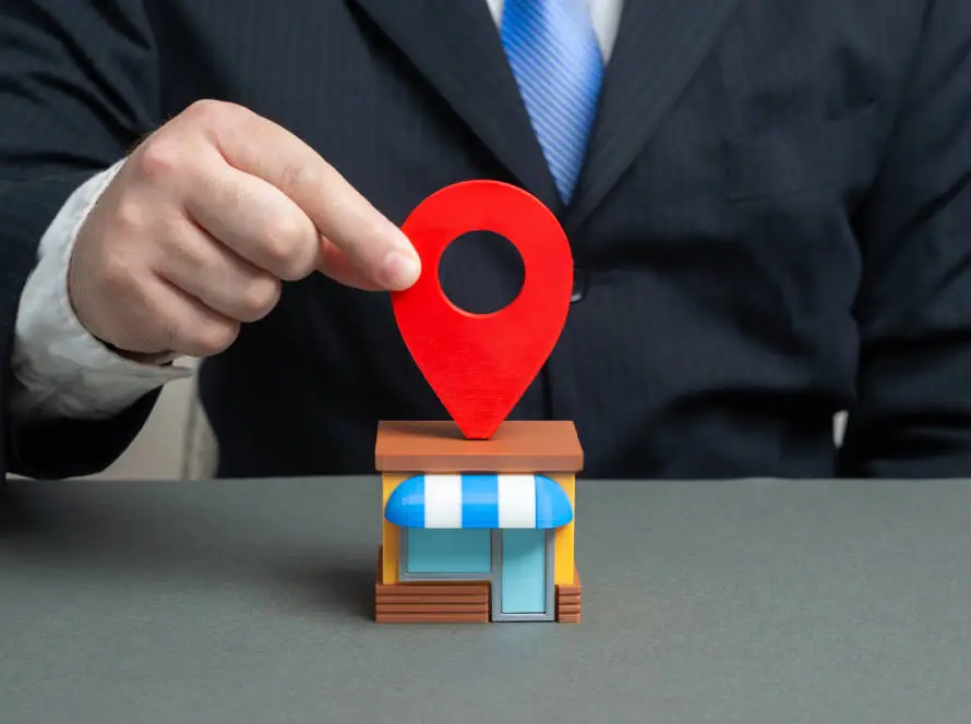 A person in a dark suit and blue tie places a large red map location pin on a model shop with a blue-and-white striped awning, symbolising local business presence—ideal for showcasing digital marketing healthcare or healthcare SEO strategies.