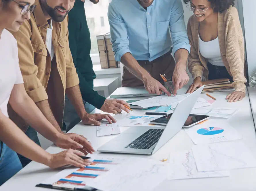 Five people stand around a table covered with charts and a laptop, collaborating and sharing ideas for a healthcare digital marketing agency in a bright, modern office setting, engaged and smiling as they shape new strategies.