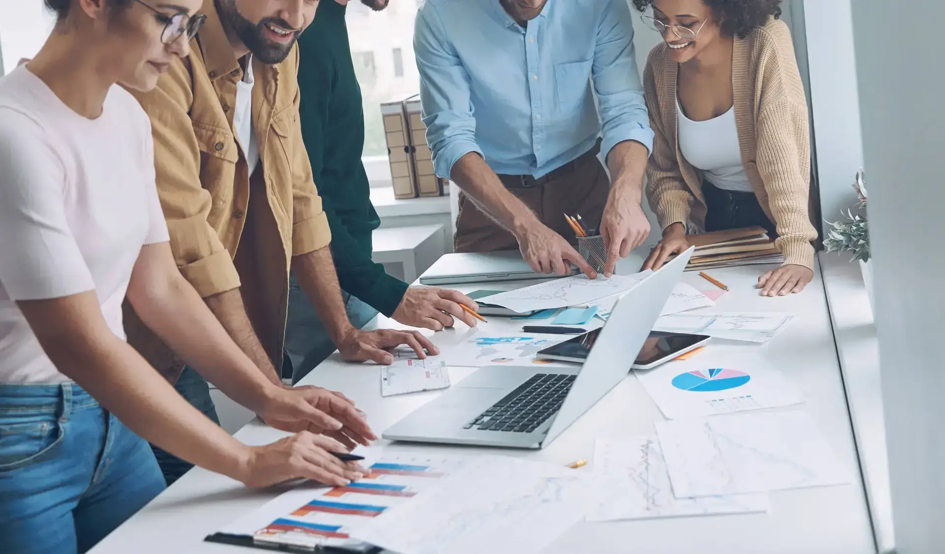 Five people stand around a table covered with charts and a laptop, collaborating and sharing ideas for a healthcare digital marketing agency in a bright, modern office setting, engaged and smiling as they shape new strategies.