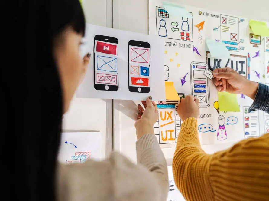 Two people collaborate on mobile app design, pinning sketches and colourful UX diagrams to a wall filled with notes and charts, illustrating a creative design process led by a healthcare digital marketing agency focused on user experience.