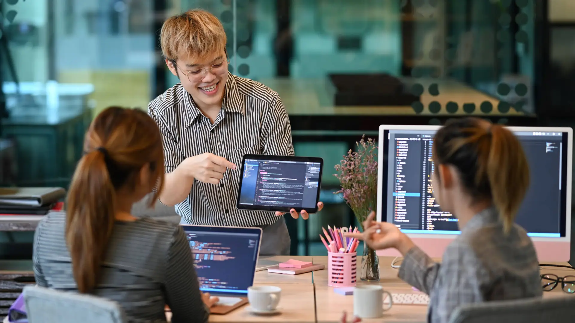 Three people sit at a table with laptops and monitors showing code. One stands, smiling and pointing at a tablet displaying code, engaging the group—a healthcare digital marketing agency team brainstorming in a modern office with pink stationery.