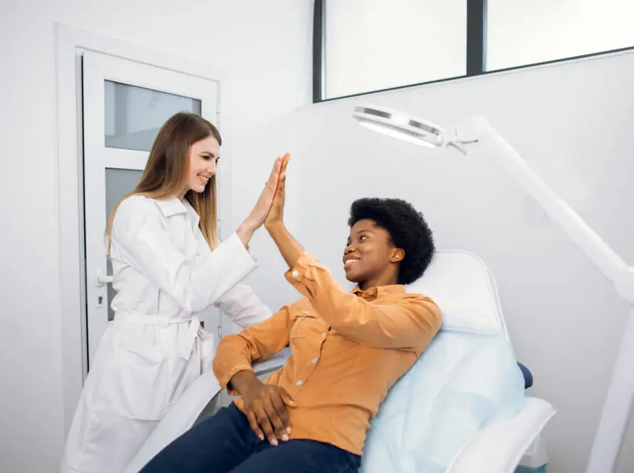 A smiling healthcare professional in a white coat gives a high five to a happy patient in an orange shirt at a modern clinic—capturing the kind of positive interactions supported by an expert medical marketing agency.