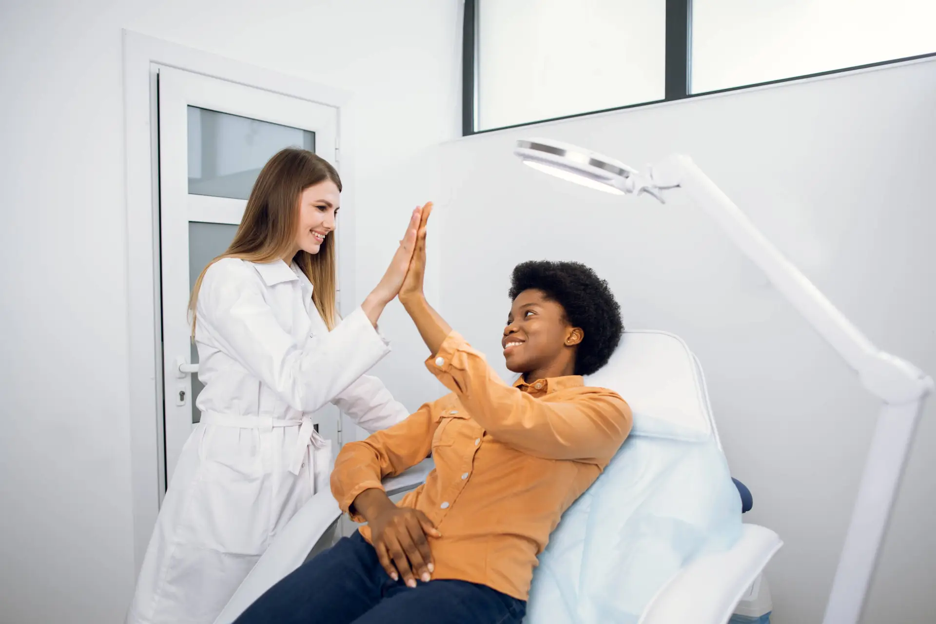 A smiling healthcare professional in a white coat gives a high five to a happy patient in an orange shirt at a modern clinic—capturing the kind of positive interactions supported by an expert medical marketing agency.