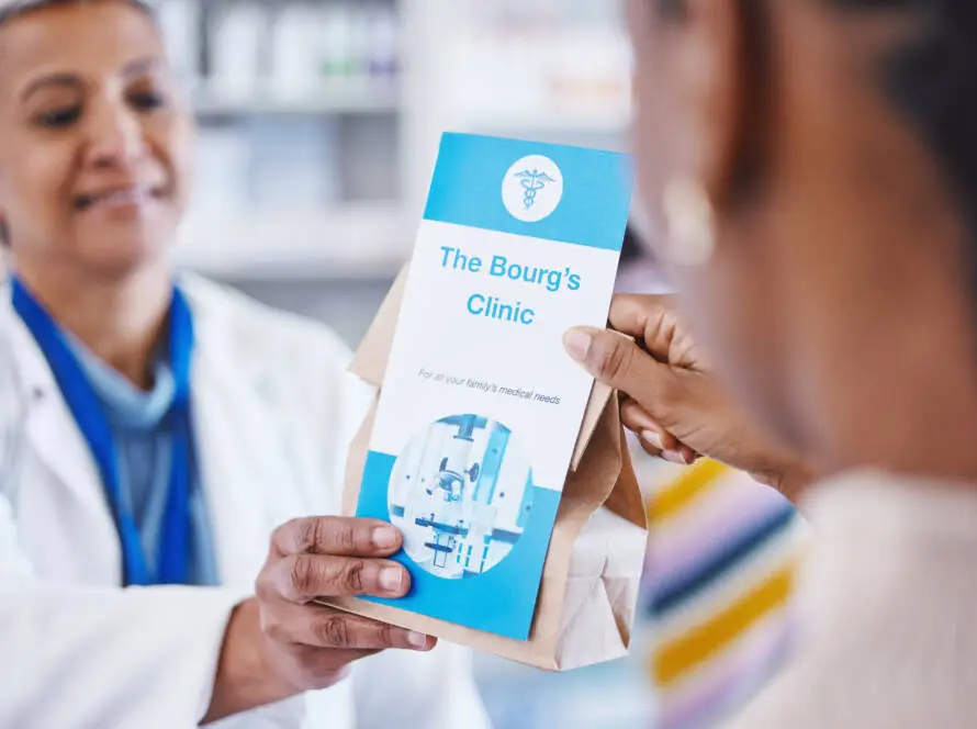 A pharmacist hands a paper bag and a clinic brochure titled “The Bourg’s Clinic” to a patient. The blue, medical-symbol brochure reflects the clinic’s partnership with a healthcare digital marketing agency. Pharmacy shelves are blurred in the background.