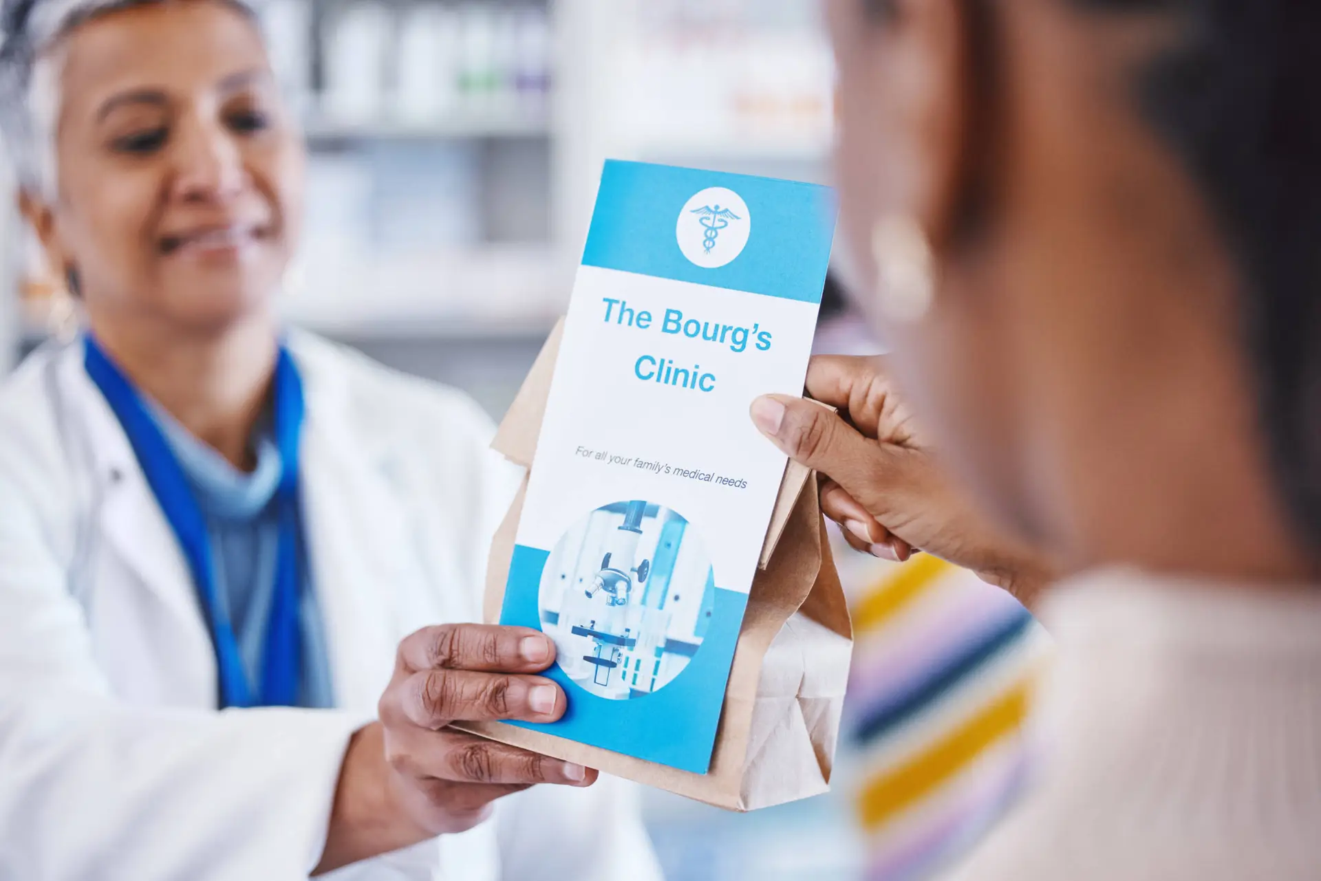 A pharmacist hands a paper bag and a clinic brochure titled “The Bourg’s Clinic” to a patient. The blue, medical-symbol brochure reflects the clinic’s partnership with a healthcare digital marketing agency. Pharmacy shelves are blurred in the background.
