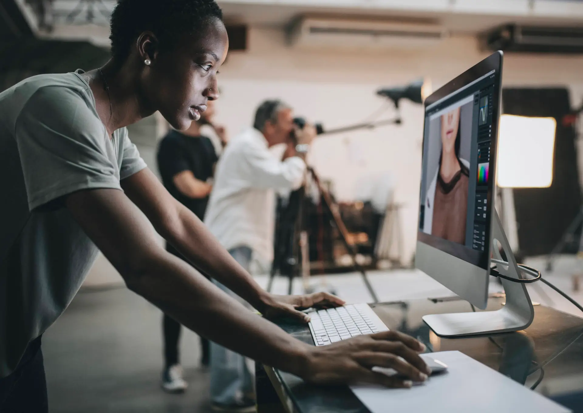 A person edits a portrait photo on a desktop computer in a photography studio. In the background, photographers with cameras and lighting equipment capture images—a modern, creative space ideal for a digital marketing healthcare team’s collaborative work.