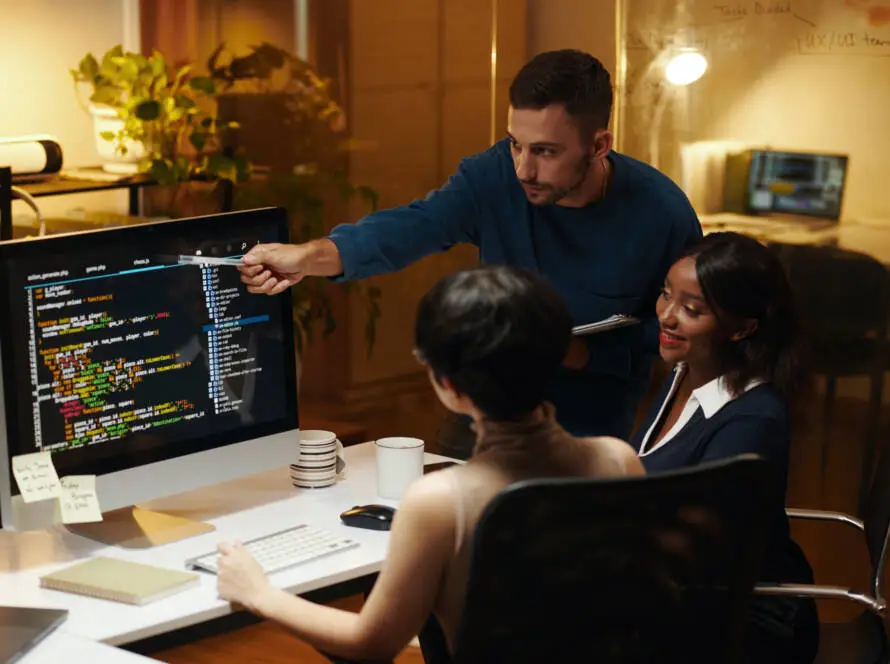 Three colleagues collaborate at a desk with a large monitor displaying code for a healthcare digital marketing agency. One stands, pointing at the screen, while two sit and focus. The office is warmly lit, with plants and laptops in the background.