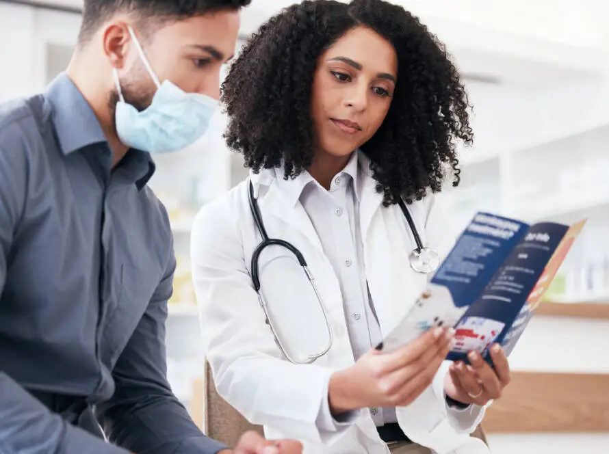 A female doctor with curly hair and a stethoscope shows a leaflet to a male patient wearing a face mask. They are seated indoors, discussing medical information in a bright, modern setting, much like a meeting with an NHS SEO consultant.