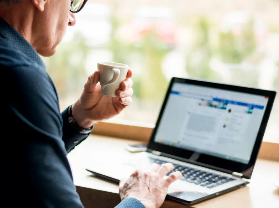 A man with grey hair and glasses sits at a wooden table, holding a small white cup in his right hand whilst using his laptop to browse a healthcare digital marketing agency website in a bright, softly blurred indoor setting.