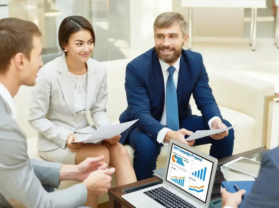 Four business professionals sit in a meeting, holding papers and smiling. A laptop on the table displays graphs and charts labelled “Startup Project” for a healthcare digital marketing agency. The atmosphere is collaborative, with everyone dressed in formal business attire.