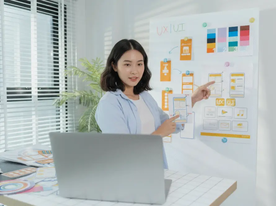 A woman stands next to a whiteboard with colourful UX/UI design sketches and colour swatches, pointing at mobile app wireframes for a digital marketing healthcare project. She faces a laptop on a desk covered with design materials as sunlight streams through window blinds.