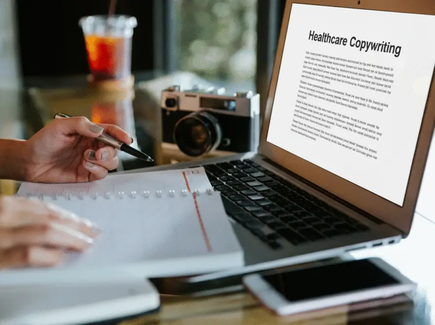 A person writes in a notebook while referencing a laptop screen displaying an article titled “Healthcare Copywriting.” Tools like a mobile phone, camera, and iced drink rest on the desk—perfect for someone working at a digital marketing healthcare agency.