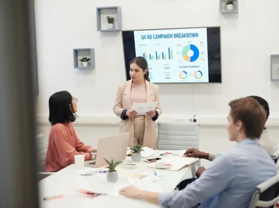 A woman in a beige blazer presents papers to three colleagues in a modern office of a medical marketing agency. Behind her, a screen displays colourful charts titled “Q4 Ad Campaign Breakdown,” with small potted plants decorating the white walls.