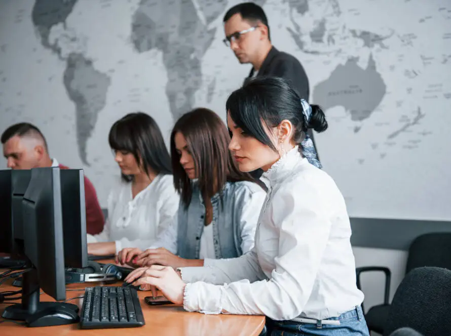 Five adults work at computers in a modern classroom with a world map on the wall. The collaborative, professional atmosphere is ideal for a medical marketing agency team focused on digital marketing healthcare strategies.