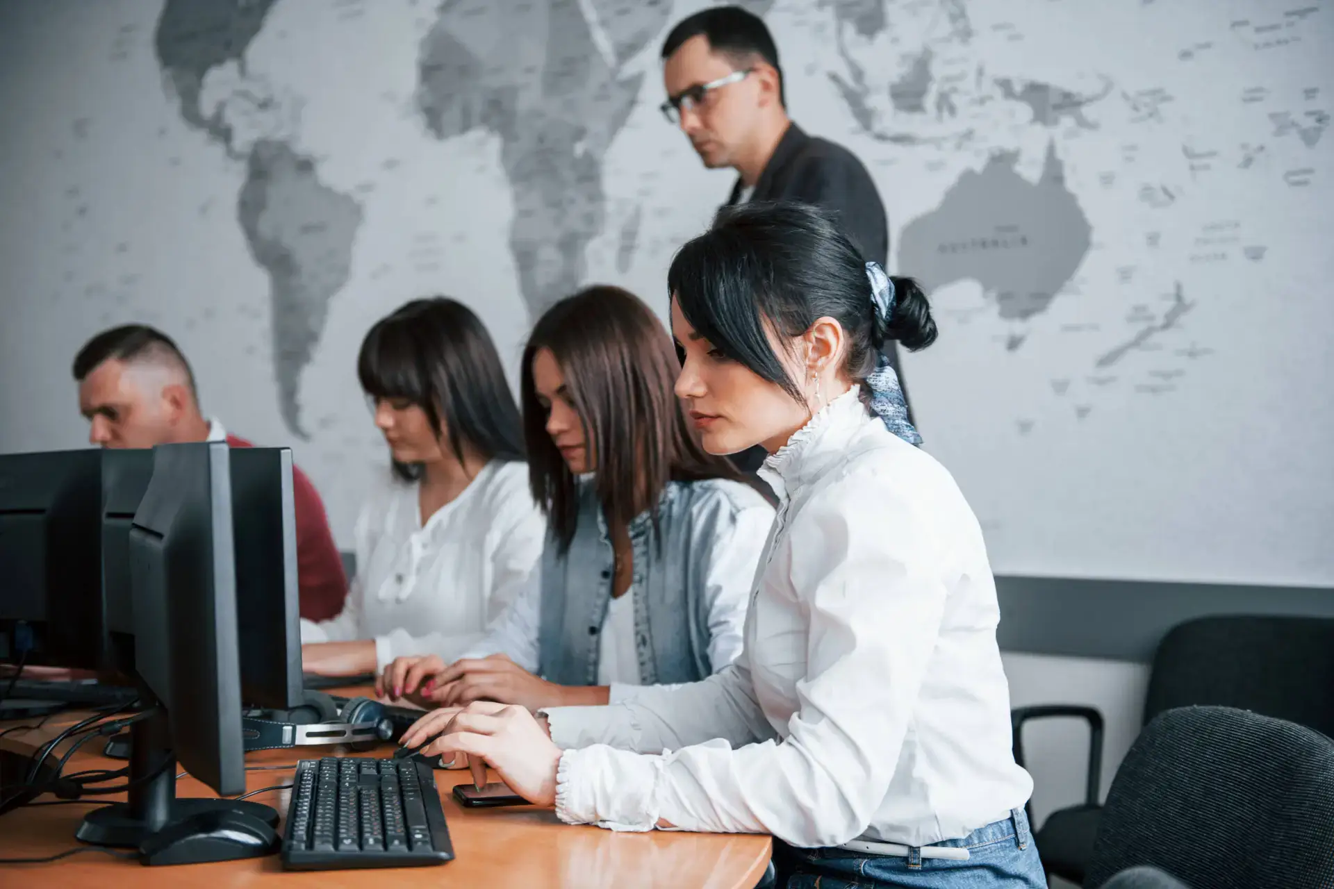 Five adults work at computers in a modern classroom with a world map on the wall. The collaborative, professional atmosphere is ideal for a medical marketing agency team focused on digital marketing healthcare strategies.