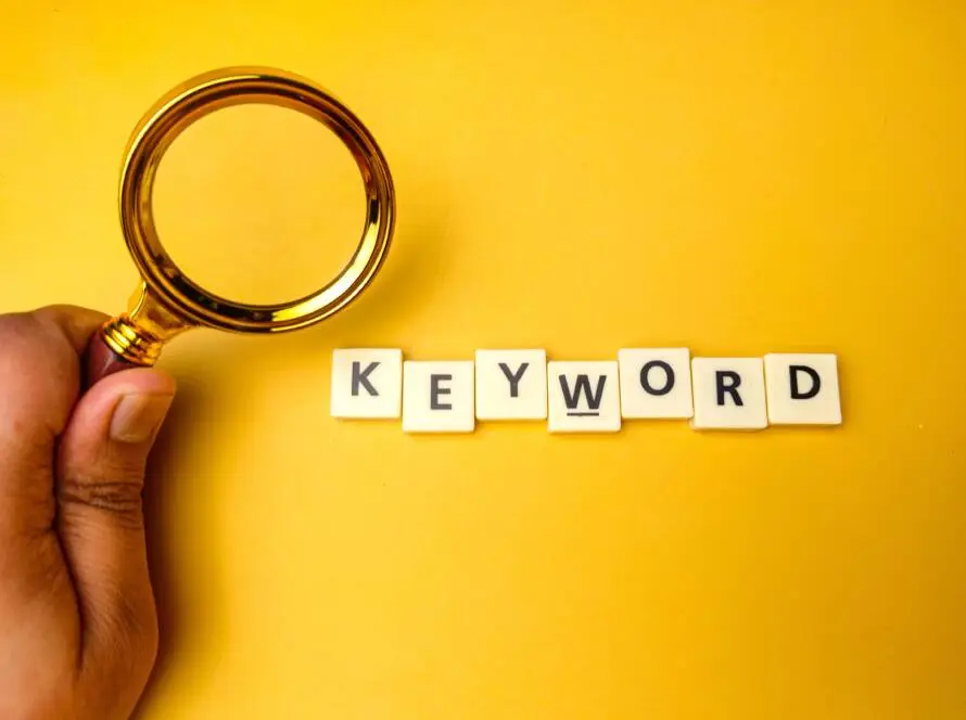 A hand holding a gold magnifying glass hovers over white letter tiles spelling “KEYWORD” on a bright yellow background—a perfect visual for a healthcare SEO agency focussed on keyword strategy.