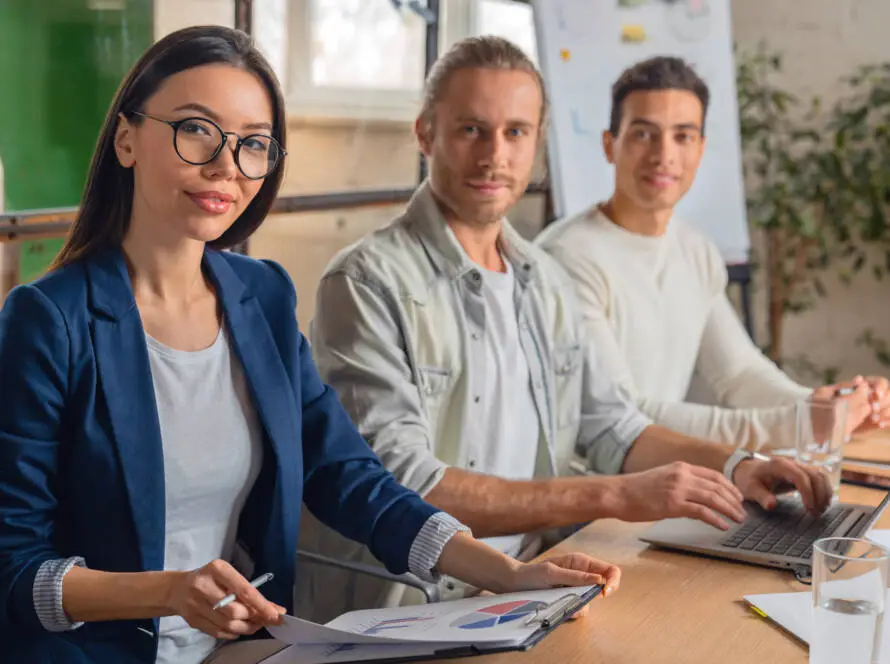 Three young professionals sit at a table in a bright office. A woman in glasses and a blue blazer holds papers. Representing a healthcare digital marketing agency, they smile at the camera with charts, documents, and water glasses on the table.