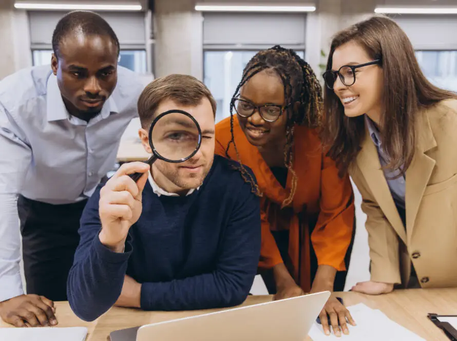 Four colleagues gather around a desk, smiling and looking at a laptop. The man in front, wearing a navy jumper, holds a magnifying glass to his eye—showcasing teamwork and the curious spirit of a healthcare SEO consultant office setting.