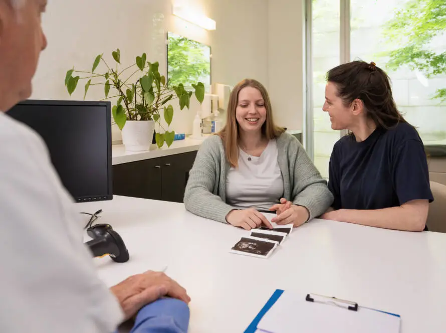 Two women sit at a desk in a bright medical office, smiling and holding hands whilst looking at ultrasound images. A healthcare digital marketing agency could capture such heartfelt moments to enhance patient engagement online.