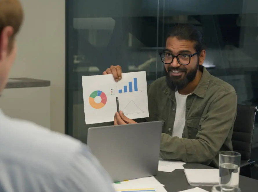 A man with a beard and glasses, wearing a green jacket, sits at a desk with a laptop. He smiles and shows a printed chart to another person, discussing results for their healthcare digital marketing agency. Papers and a glass of water are on the desk.