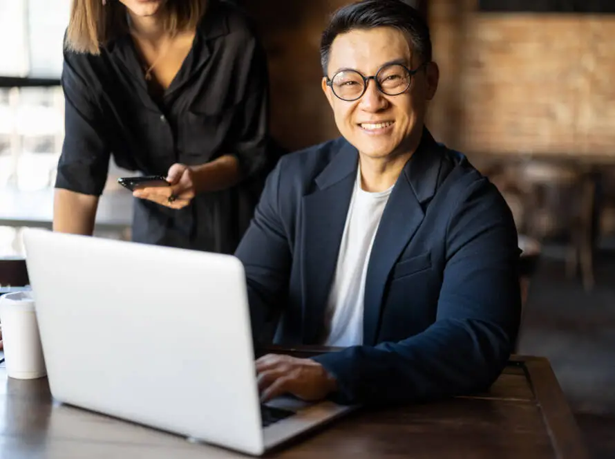 A smiling man in glasses and a dark blazer sits at a table with a laptop, working on healthcare SEO. A woman stands beside him, holding a mobile phone. The background is a bright, stylish café with exposed brick walls.