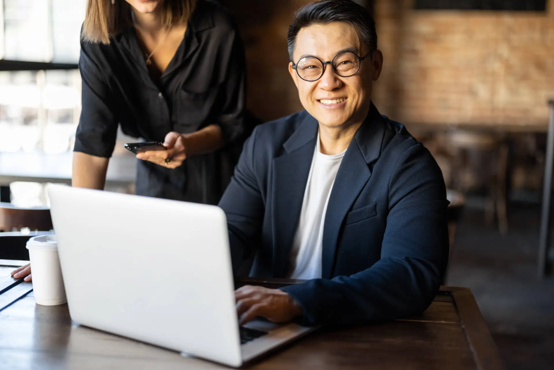 A smiling man in glasses and a dark blazer sits at a table with a laptop, working on healthcare SEO. A woman stands beside him, holding a mobile phone. The background is a bright, stylish café with exposed brick walls.